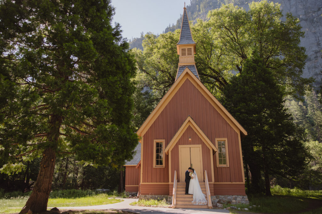 A couple dressed in wedding attire stands on the steps of a small, brown chapel surrounded by trees at Yosemite Chapel for their yosemite elopement 