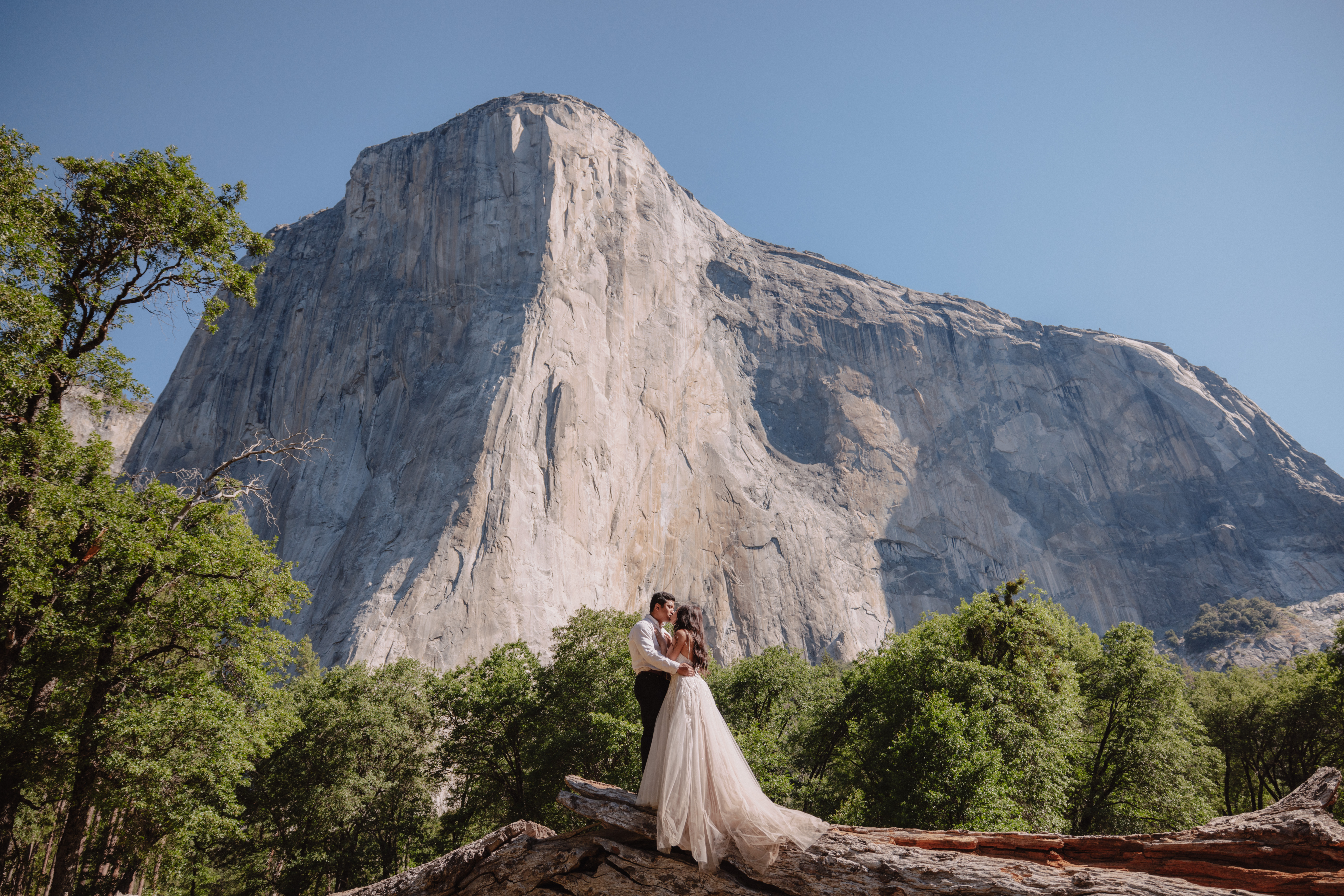 A couple stands in an embrace on a large rock amidst greenery with a towering cliff face in the background under a clear blue sky.