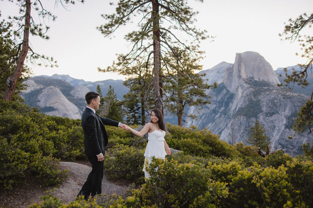 A couple in formal attire walking hand-in-hand on a nature trail with mountains and trees in the background for their Yosemite elopement