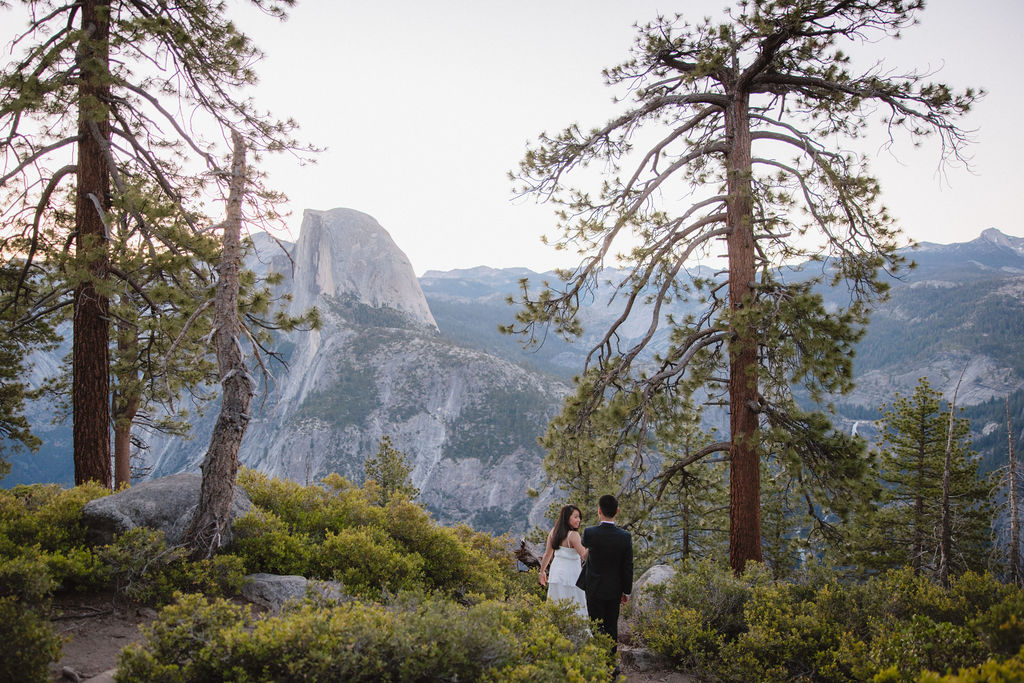 A couple in formal attire walking hand-in-hand on a nature trail with mountains and trees in the background for their Yosemite elopement