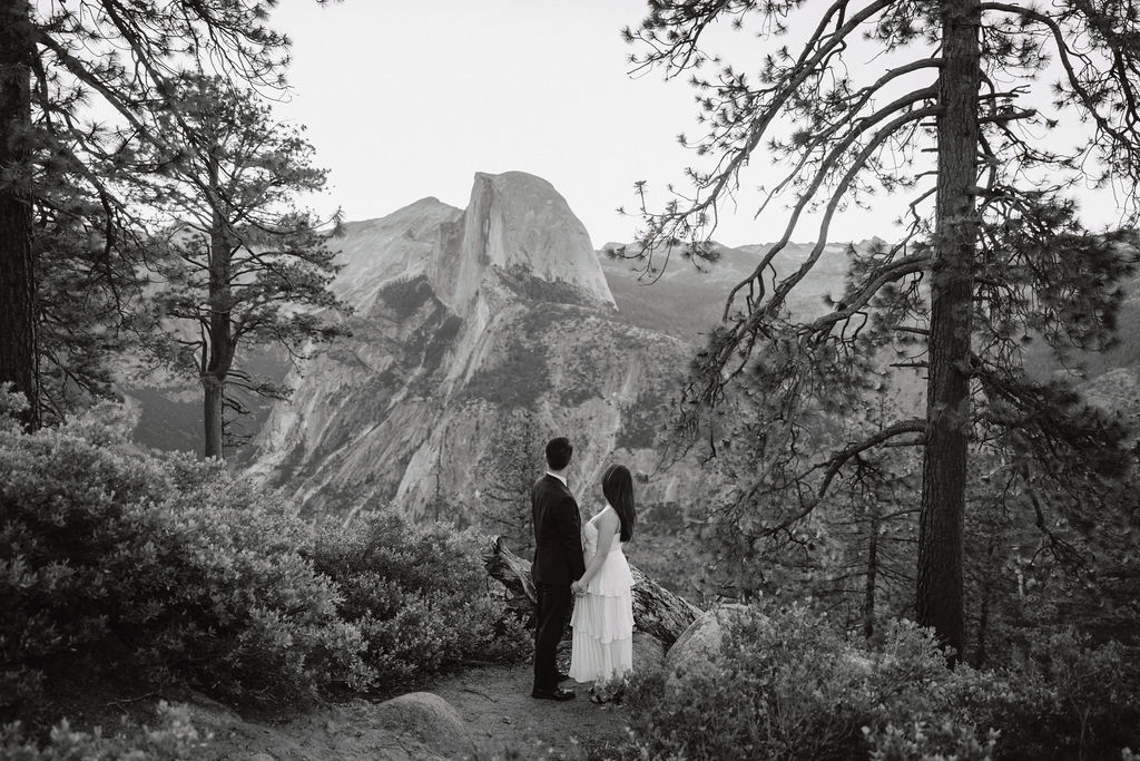 A couple in formal attire walking hand-in-hand on a nature trail with mountains and trees in the background for their Yosemite elopement