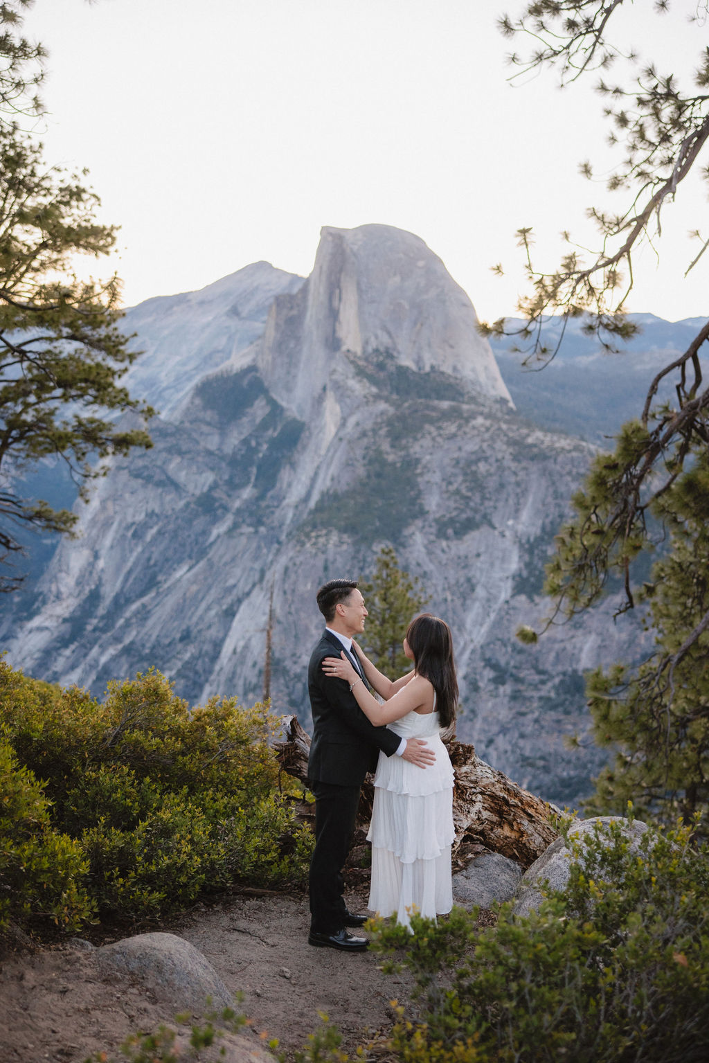 A couple in formal attire walking hand-in-hand on a nature trail with mountains and trees in the background for their Yosemite elopement