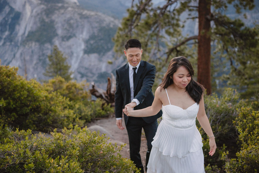 A couple in formal attire walking hand-in-hand on a nature trail with mountains and trees in the background for their Yosemite elopement