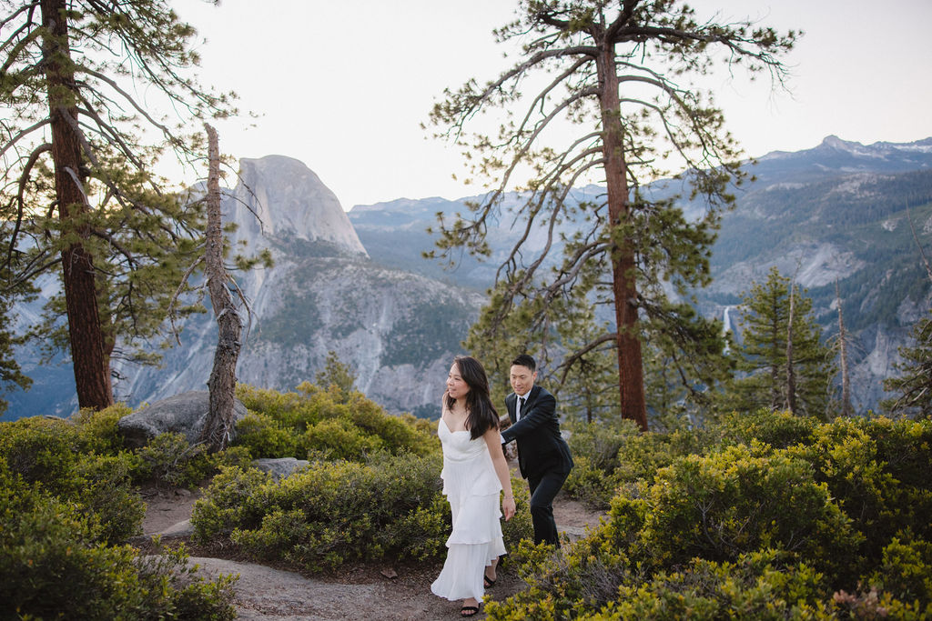 A couple in formal attire walking hand-in-hand on a nature trail with mountains and trees in the background for their Yosemite elopement