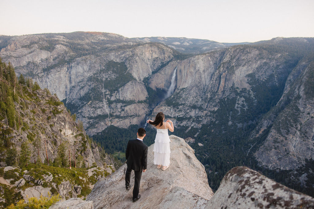 A couple, dressed in black and white, stands on a cliff edge overlooking a vast mountainous landscape, with trees and rocky cliffs in the background for their Yosemite elopement