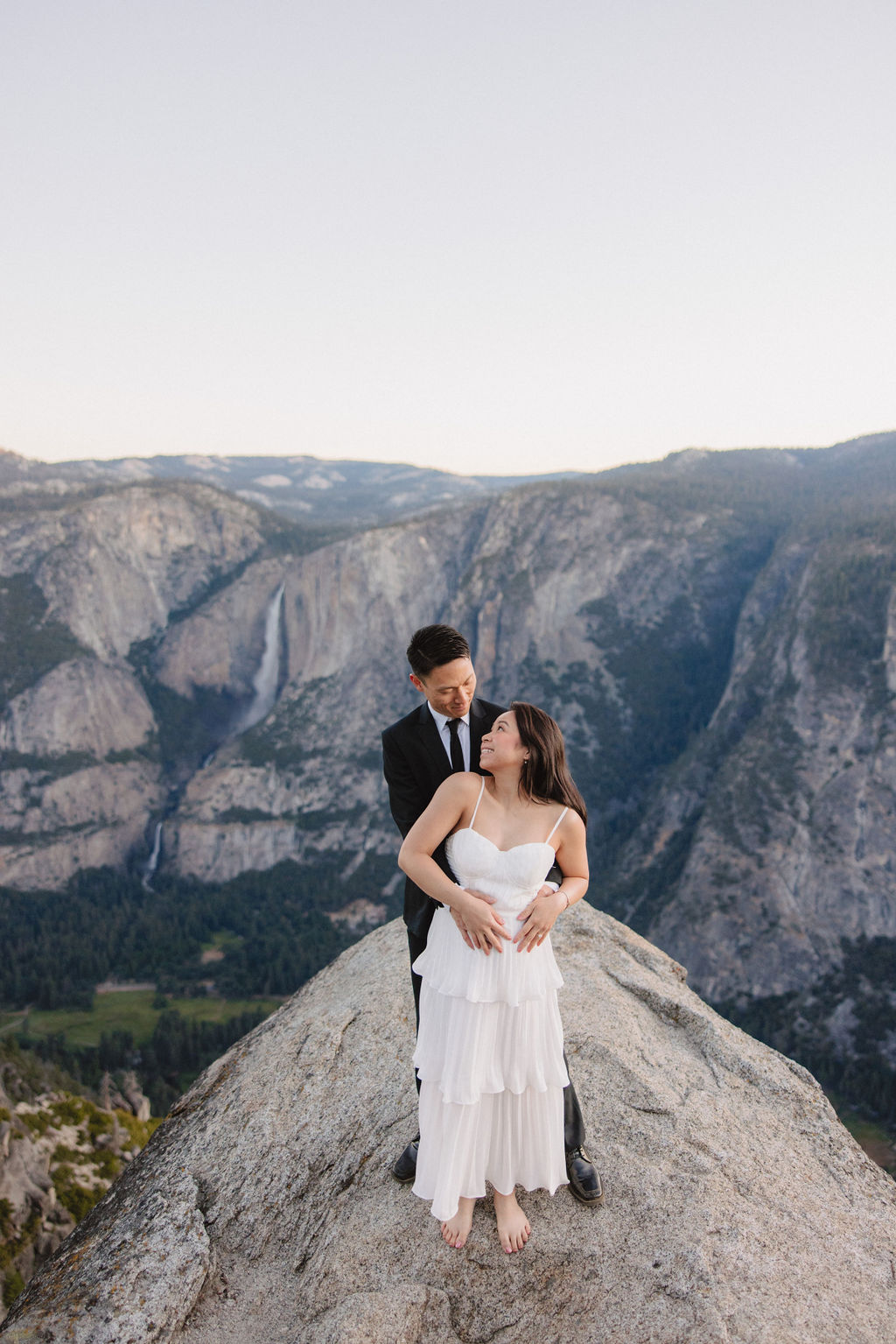 A couple, dressed in black and white, stands on a cliff edge overlooking a vast mountainous landscape, with trees and rocky cliffs in the background for their Yosemite elopement