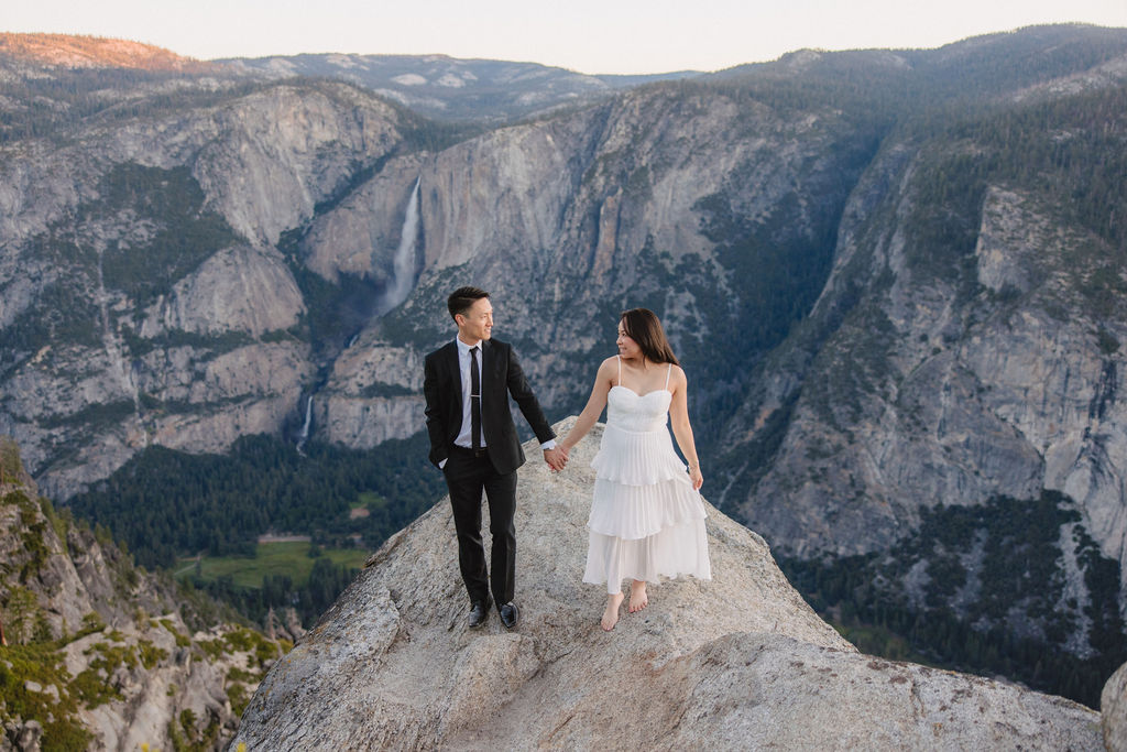 A couple, dressed in black and white, stands on a cliff edge overlooking a vast mountainous landscape, with trees and rocky cliffs in the background for their Yosemite elopement