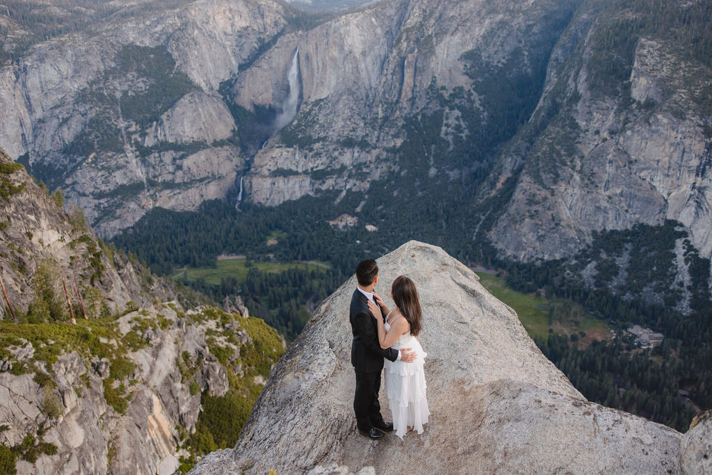 A couple, dressed in black and white, stands on a cliff edge overlooking a vast mountainous landscape, with trees and rocky cliffs in the background for their Yosemite elopement