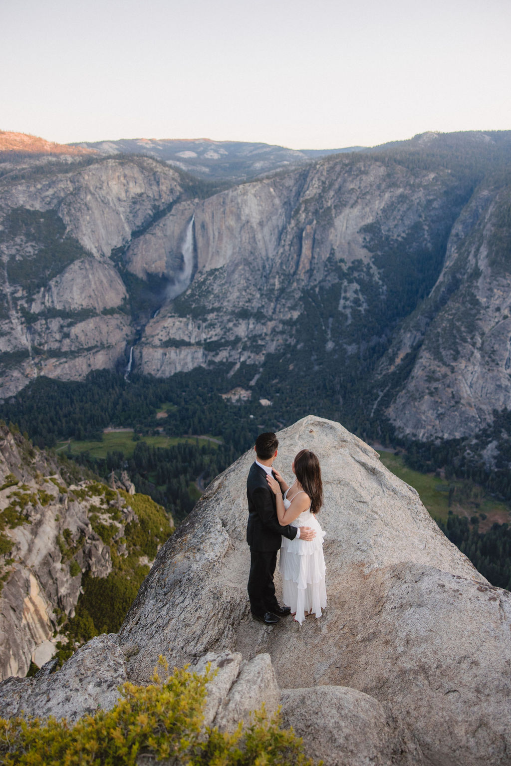 A couple, dressed in black and white, stands on a cliff edge overlooking a vast mountainous landscape, with trees and rocky cliffs in the background for their Yosemite elopement