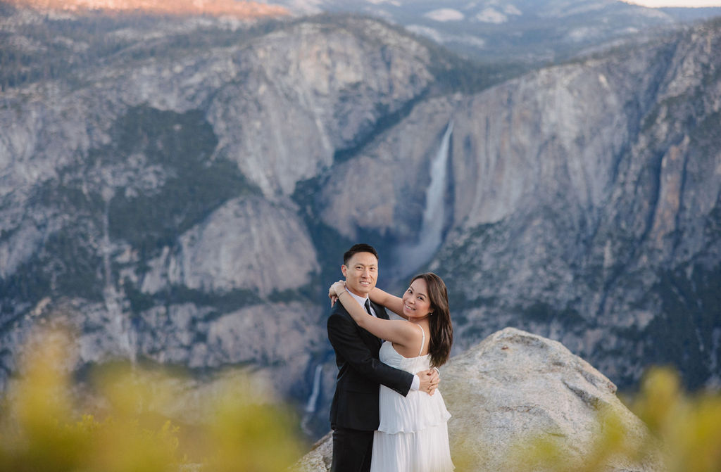 A couple, dressed in black and white, stands on a cliff edge overlooking a vast mountainous landscape, with trees and rocky cliffs in the background for their Yosemite elopement