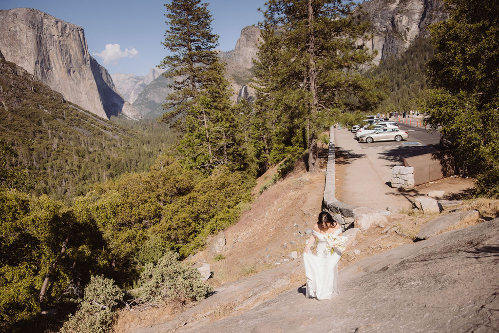 Bride stands on a rocky slope surrounded by trees and mountains, with a parking lot and cars visible in the background at Yosemite National Park