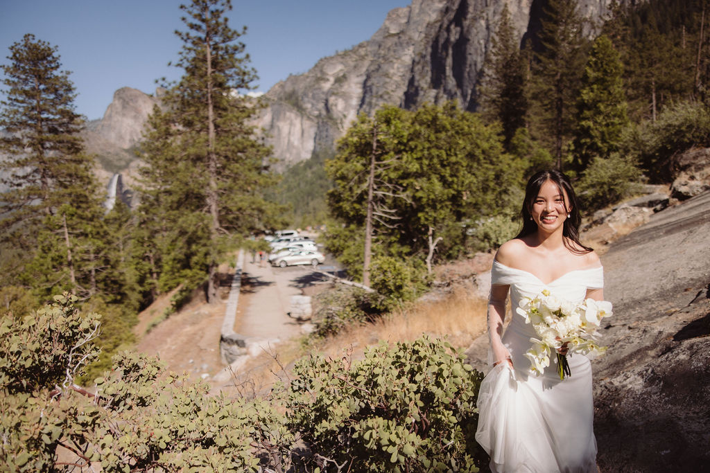 Bride stands on a rocky slope surrounded by trees and mountains, with a parking lot and cars visible in the background at Yosemite National Park