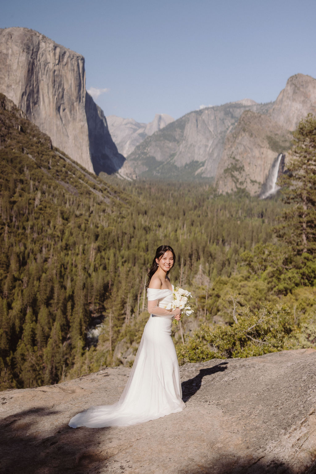 A person in a white dress stands on a rock with a bouquet, overlooking a scenic landscape of mountains, trees, and a waterfall.