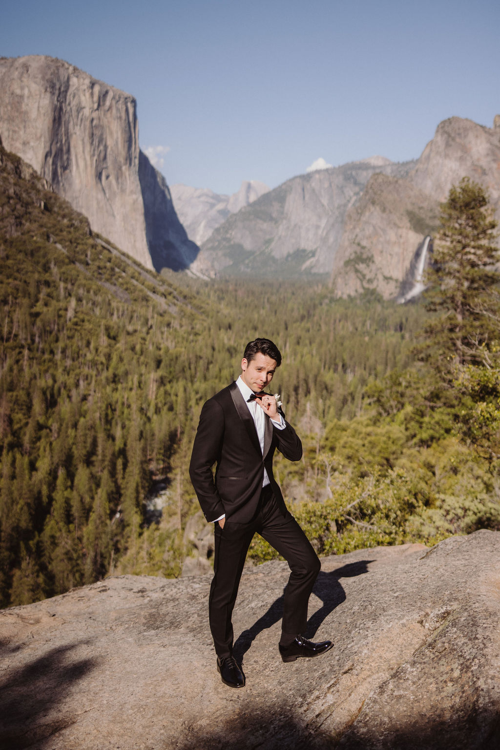 A man in a black suit stands on a rocky edge overlooking a vast, mountainous landscape with clear skies.