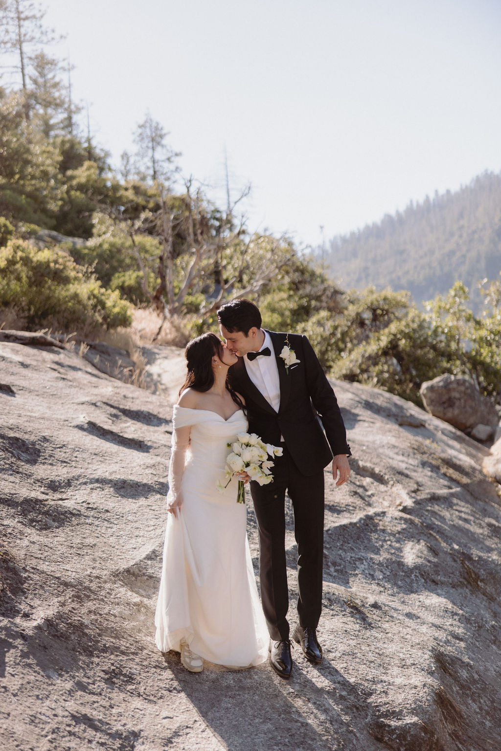 A bride in a white dress and a groom in a black suit share a kiss while standing on a rocky terrain with trees and mountains in the background during their yosemite elopment