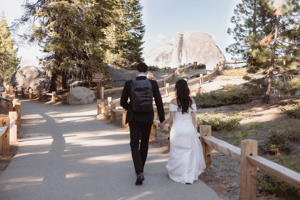 A couple, wearing formal attire, walks hand-in-hand on a paved trail through a forested area with large rocks in the background during their Taft Point Elopement 