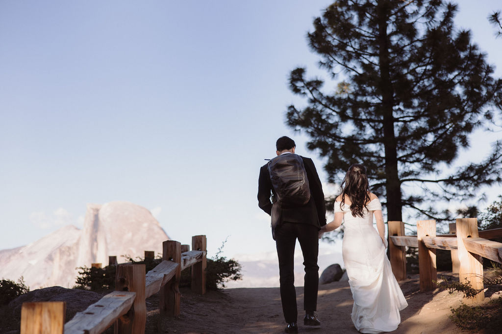 A couple, wearing formal attire, walks hand-in-hand on a paved trail through a forested area with large rocks in the background during their Taft Point Elopement 