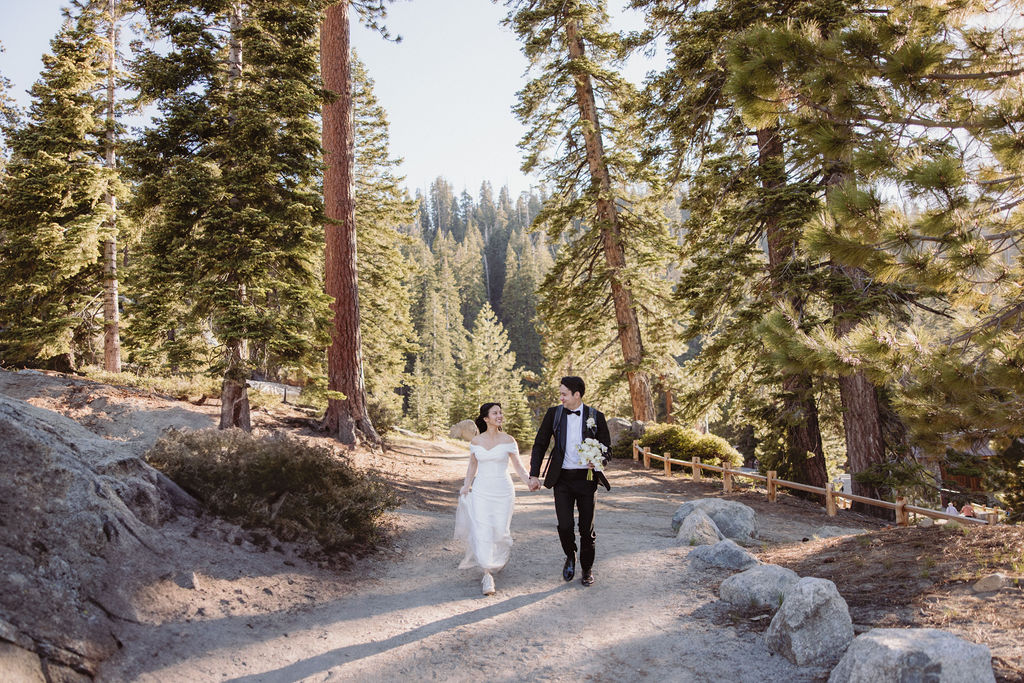 A couple in wedding attire walks hand in hand along a forested trail surrounded by tall trees and sunlight filtering through the branches during their taft point elopment at yosemite national park