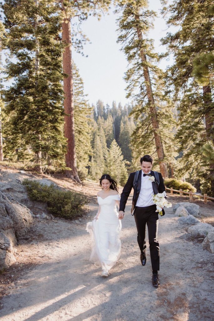 A couple in wedding attire walks hand in hand along a forested trail surrounded by tall trees and sunlight filtering through the branches during their taft point elopment at yosemite national park