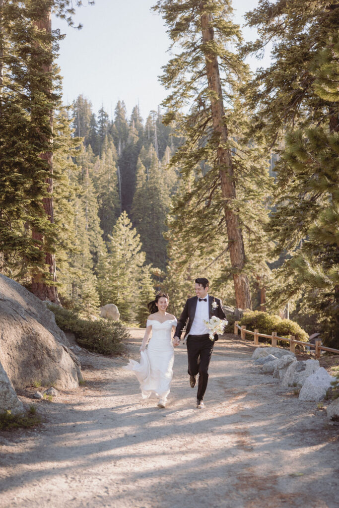 A couple in wedding attire walks hand in hand along a forested trail surrounded by tall trees and sunlight filtering through the branches during their taft point elopment at yosemite national park