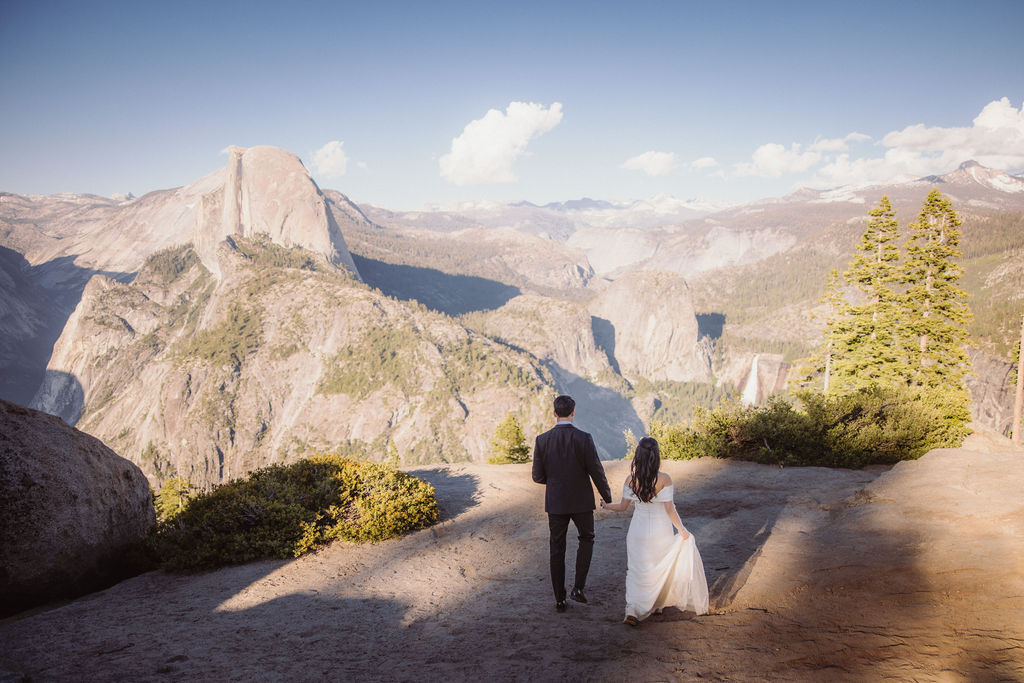 Couple in wedding attire walking hand-in-hand on a scenic mountain path with a vista of rugged mountains and trees in the background at Taft Point