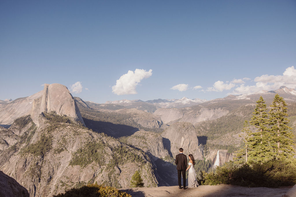 Couple in wedding attire walking hand-in-hand on a scenic mountain path with a vista of rugged mountains and trees in the background at Taft Point