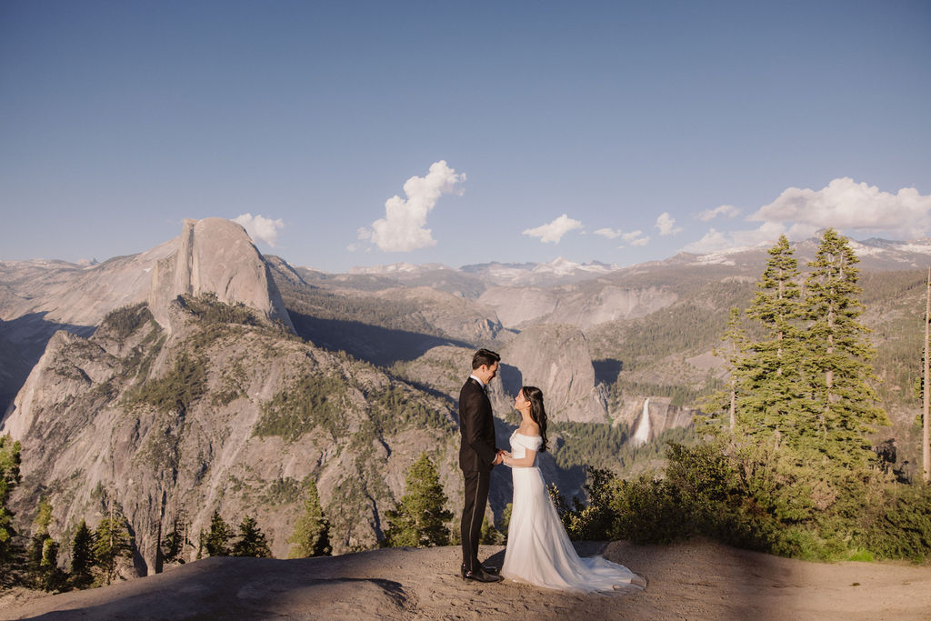 Couple in wedding attire walking hand-in-hand on a scenic mountain path with a vista of rugged mountains and trees in the background at Taft Point