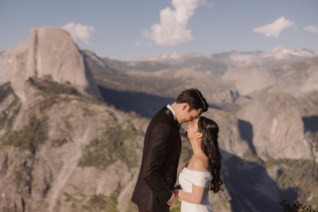 A couple wearing formal attire stands close, foreheads touching, with a scenic mountainous landscape in the background at their elopement at Taft Point