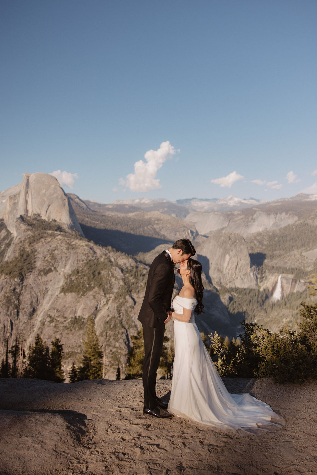 Couple in wedding attire walking hand-in-hand on a scenic mountain path with a vista of rugged mountains and trees in the background at Taft Point