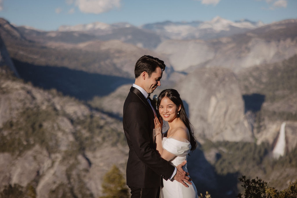 A couple wearing formal attire stands close, foreheads touching, with a scenic mountainous landscape in the background at their elopement at Taft Point