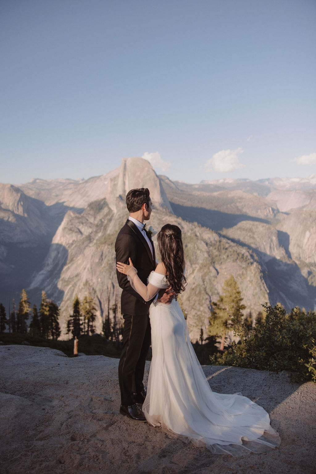 Couple in wedding attire walking hand-in-hand on a scenic mountain path with a vista of rugged mountains and trees in the background at Taft Point