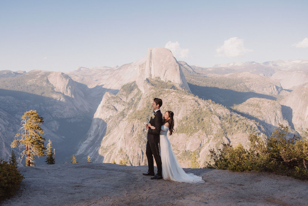 Couple in wedding attire walking hand-in-hand on a scenic mountain path with a vista of rugged mountains and trees in the background at Taft Point