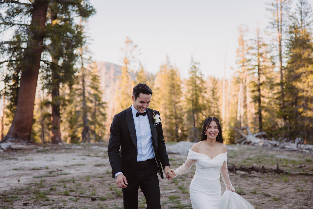 A bride and groom, dressed in a white gown and black tuxedo, walk hand in hand in a forested area during their Yosemite Elopment