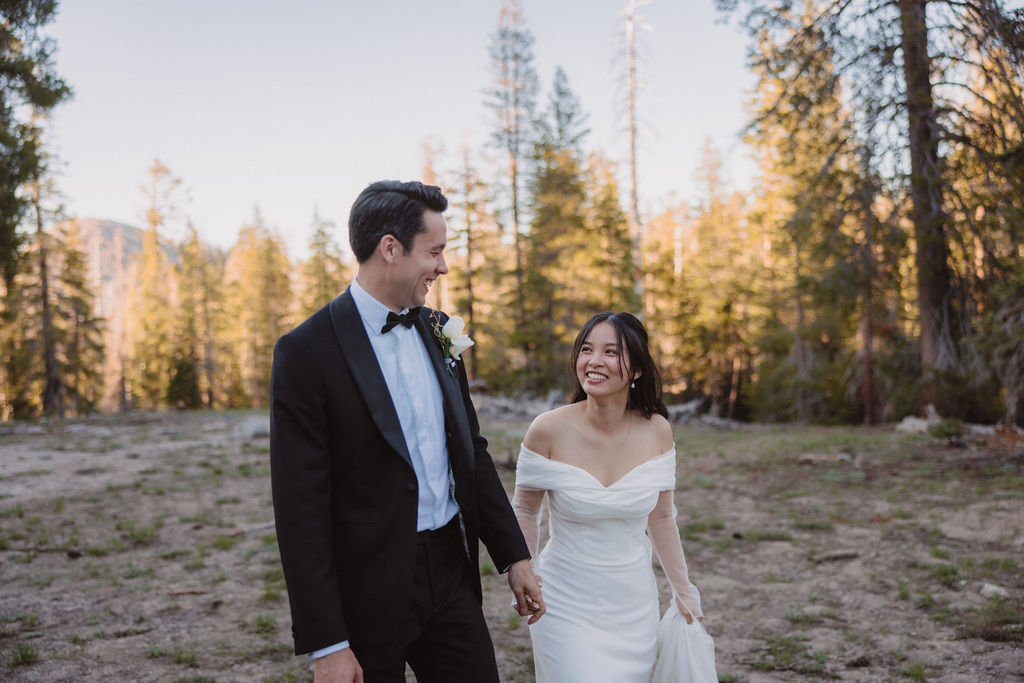 A bride and groom, dressed in a white gown and black tuxedo, walk hand in hand in a forested area during their Yosemite Elopment