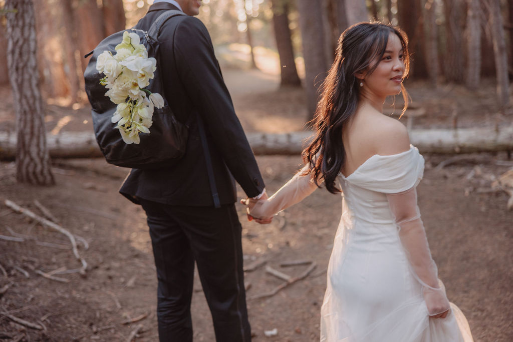 A couple, dressed in formal attire, walks hand-in-hand through a forest at sunset. The bride wears a white gown, and the groom holds a bouquet during their Yosemite elopement