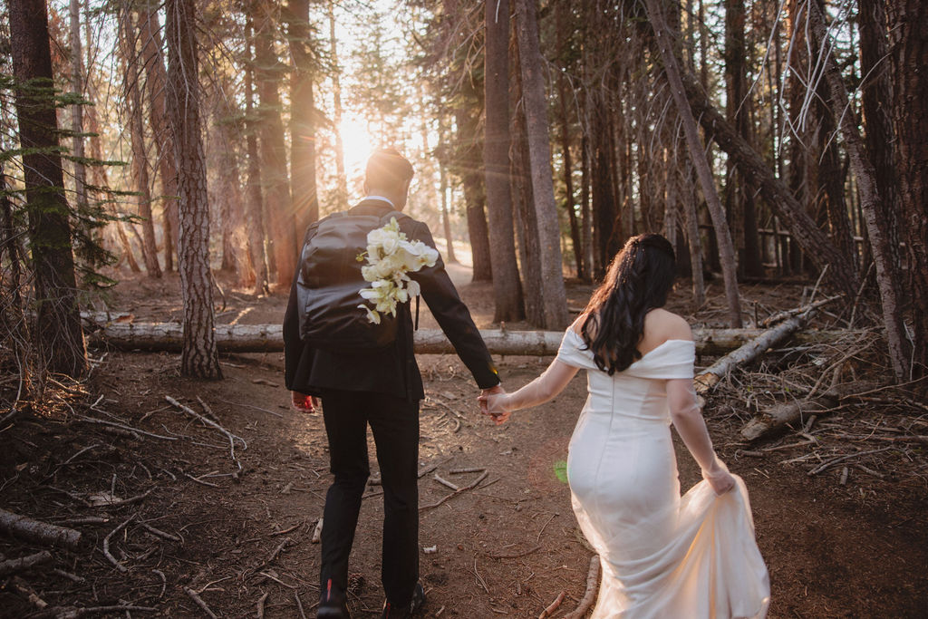 A couple, dressed in formal attire, walks hand-in-hand through a forest at sunset. The bride wears a white gown, and the groom holds a bouquet during their Yosemite elopement