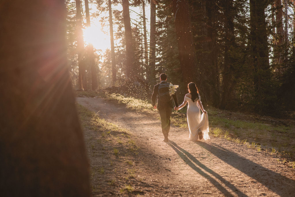 A couple, dressed in formal attire, walks hand-in-hand through a forest at sunset. The bride wears a white gown, and the groom holds a bouquet during their Yosemite elopement