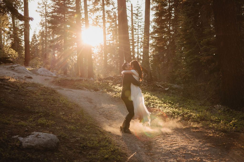 A couple, dressed in formal attire, walks hand-in-hand through a forest at sunset. The bride wears a white gown, and the groom holds a bouquet during their Yosemite elopement