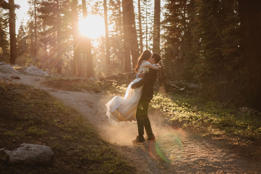 A couple, dressed in formal attire, walks hand-in-hand through a forest at sunset. The bride wears a white gown, and the groom holds a bouquet during their Yosemite elopement