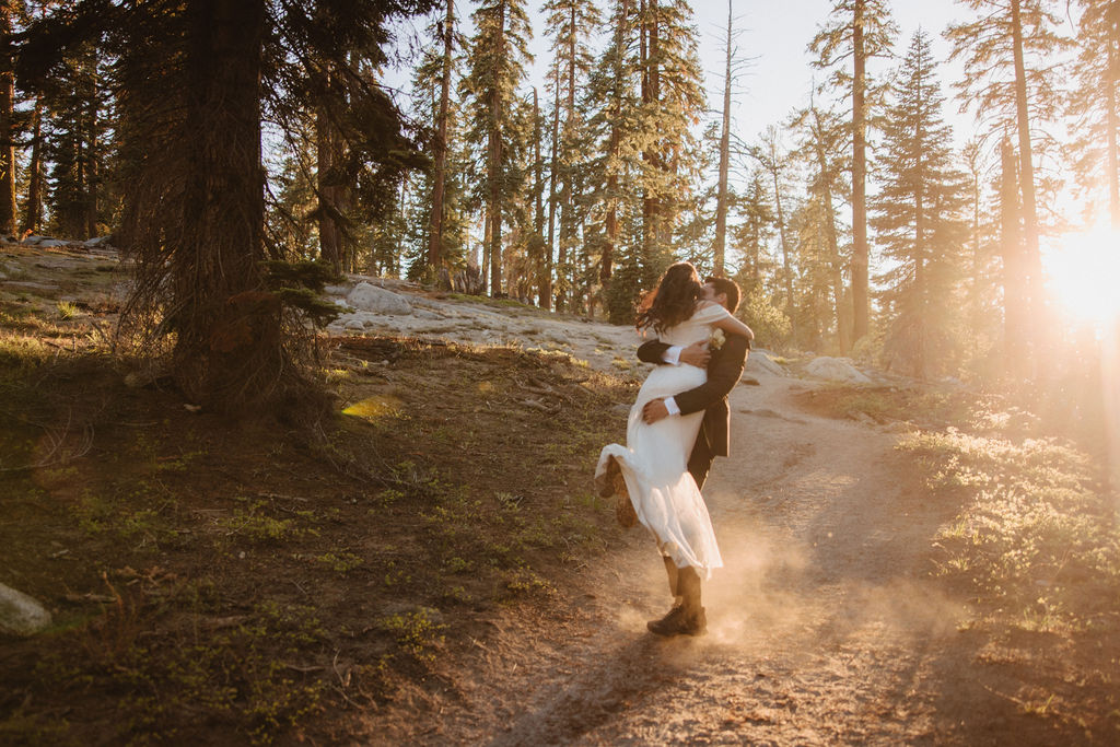 A couple, dressed in formal attire, walks hand-in-hand through a forest at sunset. The bride wears a white gown, and the groom holds a bouquet during their Yosemite elopement