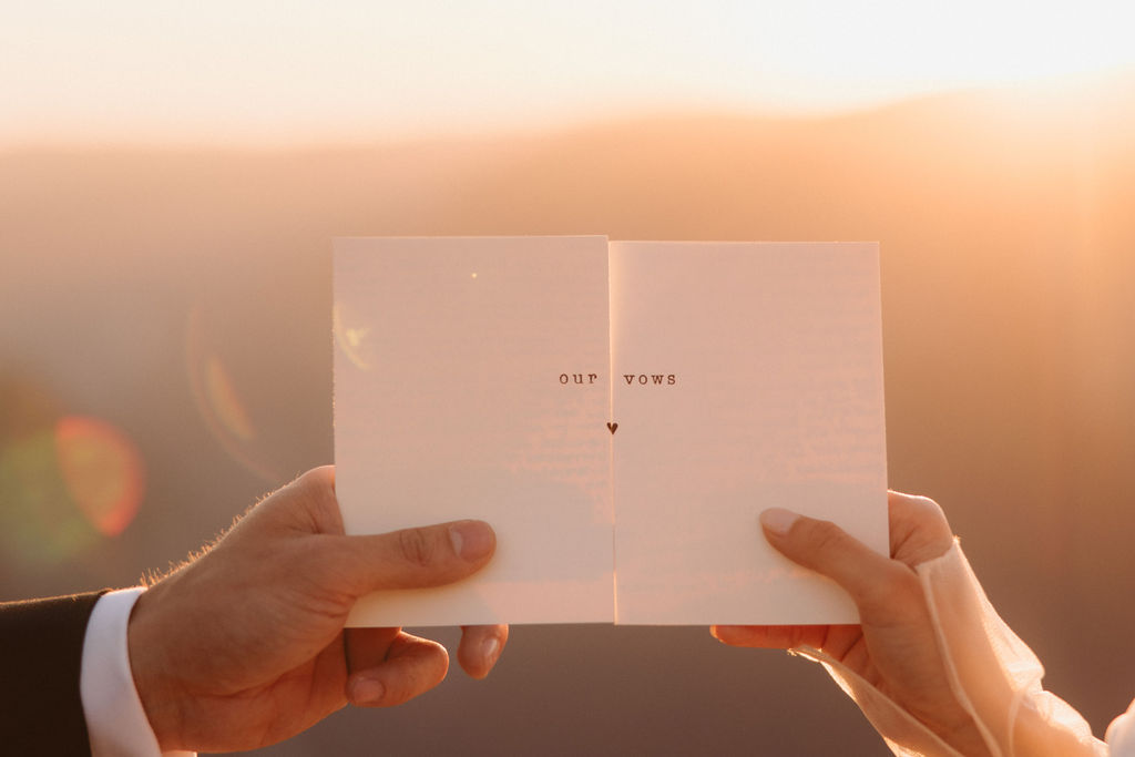 Two people holding a book open toward a sunset, each hand on opposite sides of the book. The background is warm and softly lit by the sun.