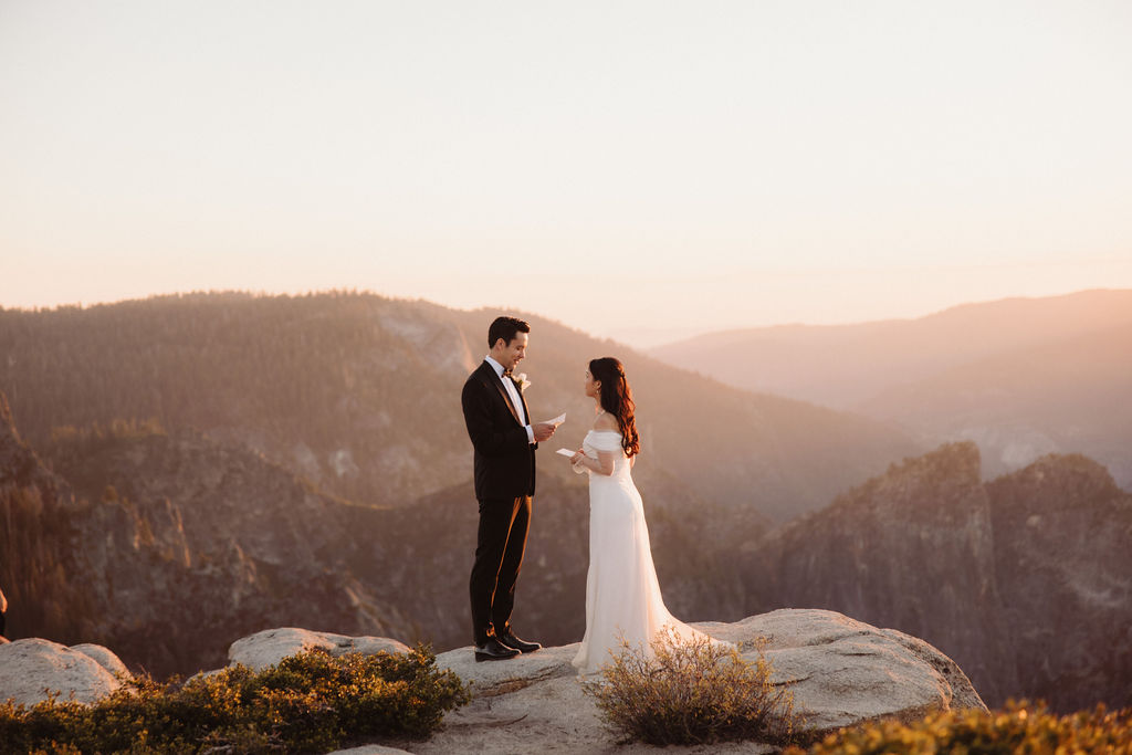 A couple stands facing each other at the edge of a cliff during sunset. The man is in a suit, and the woman is in a white dress, with mountains in the background at Taft Point 