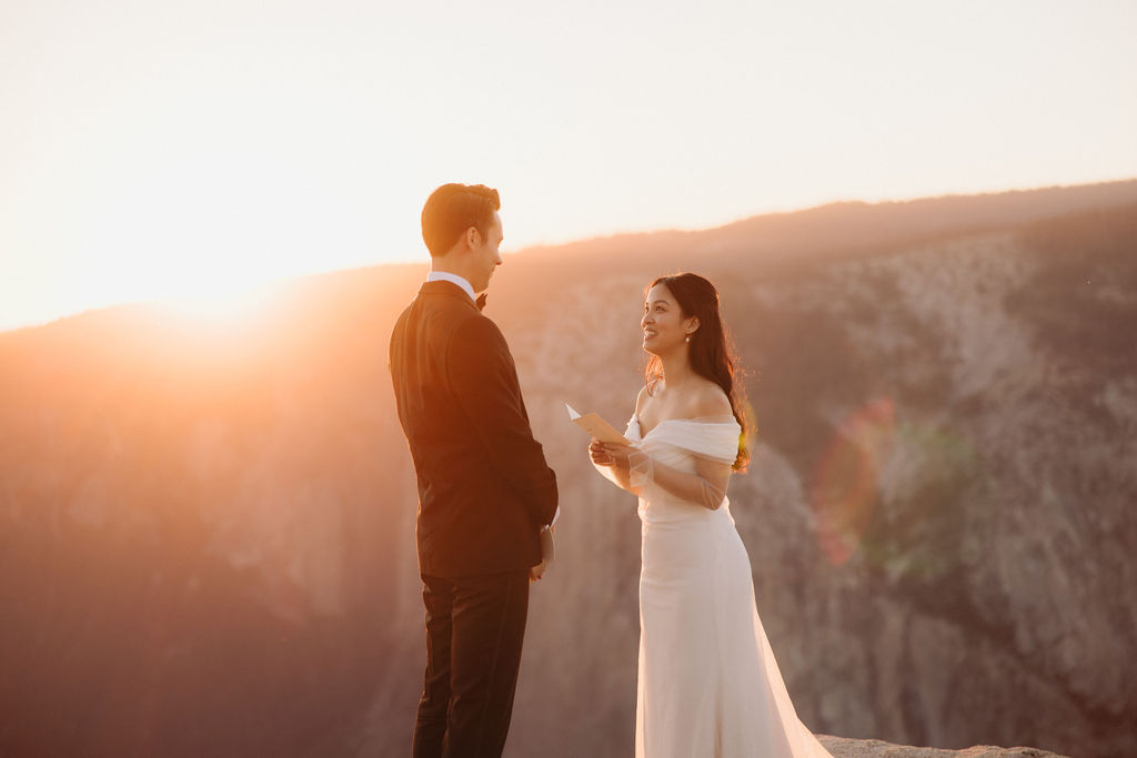 A couple stands facing each other at the edge of a cliff during sunset. The man is in a suit, and the woman is in a white dress, with mountains in the background at Taft Point 