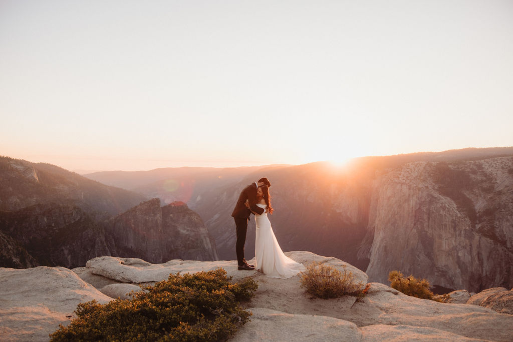 A couple stands facing each other at the edge of a cliff during sunset. The man is in a suit, and the woman is in a white dress, with mountains in the background at Taft Point