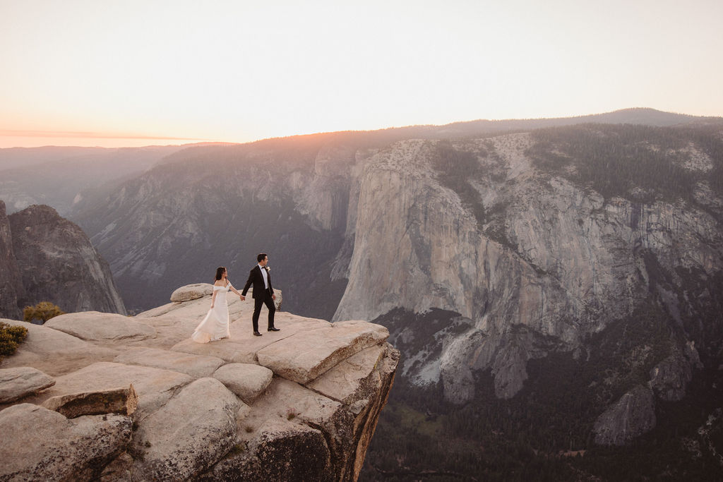 A couple in wedding attire stands on a rocky cliff edge at sunset, overlooking a vast mountainous landscape.