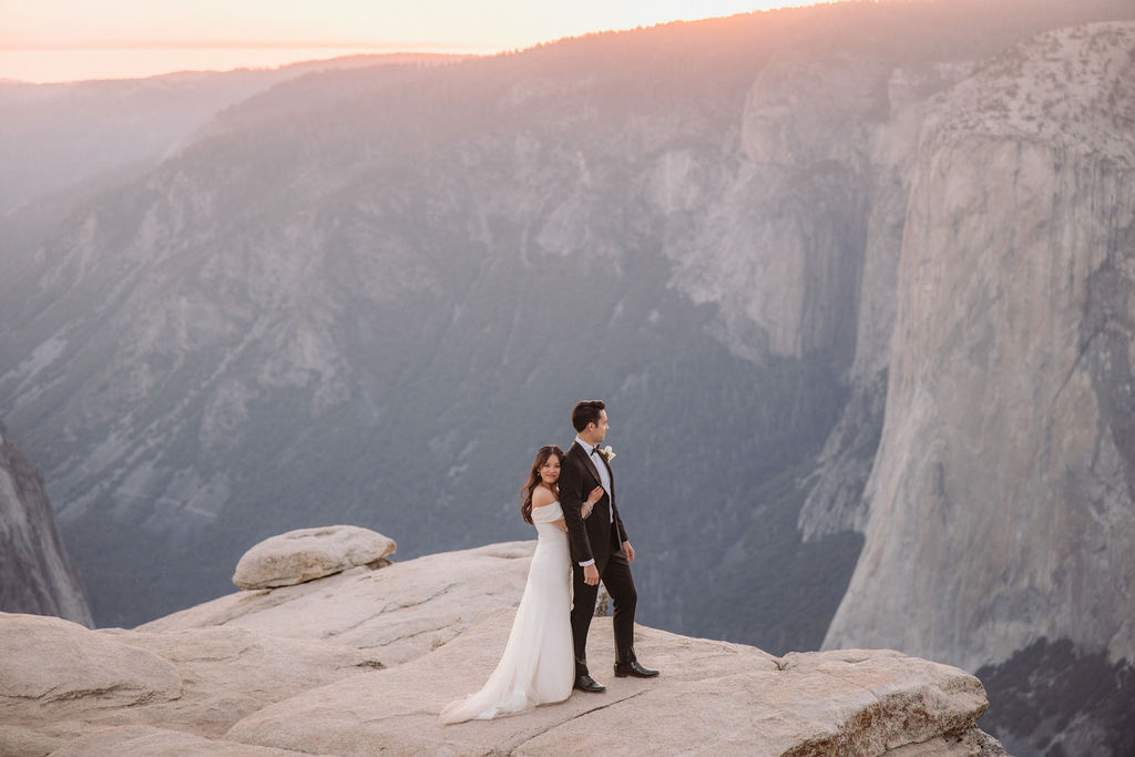 A couple dressed in wedding attire stands on a cliff edge with a vast mountainous landscape in the background at sunset during their elopement at Taft Point