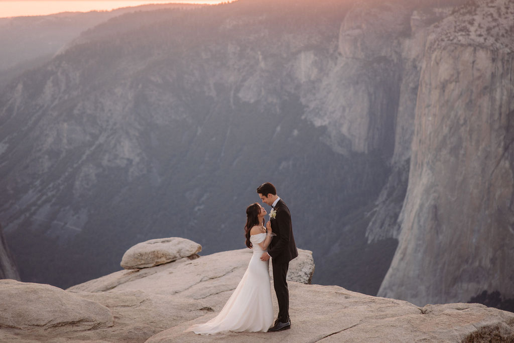 A couple in wedding attire stands on a rocky cliff edge at sunset, overlooking a vast mountainous landscape.