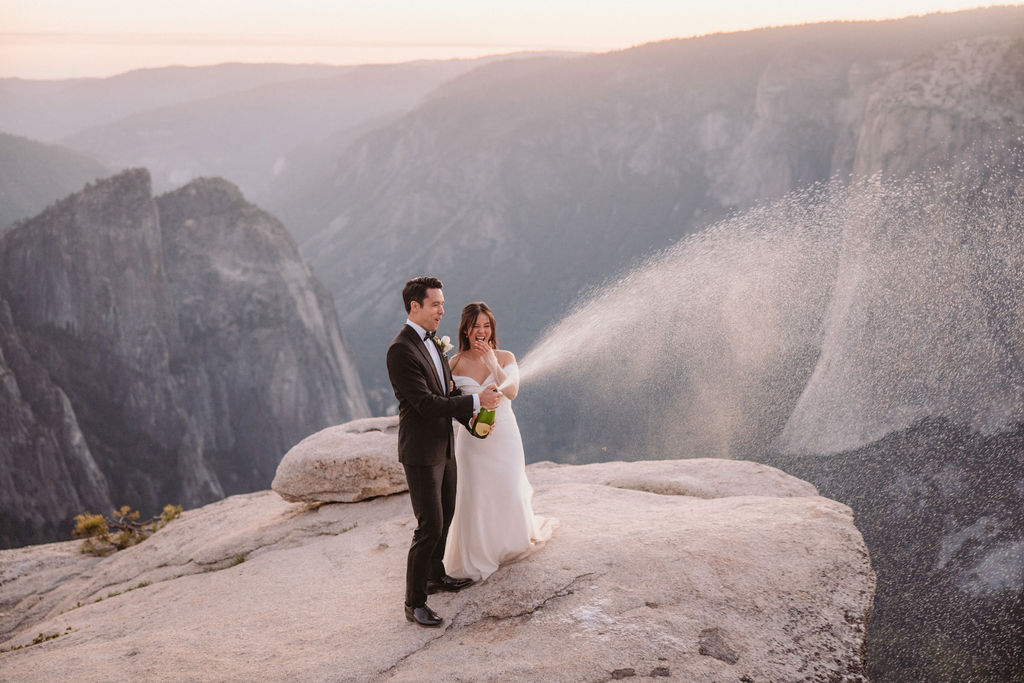 A bride and groom stand on a cliff edge, with the groom spraying champagne while the bride smiles, set against a backdrop of mountains and a sunset sky.