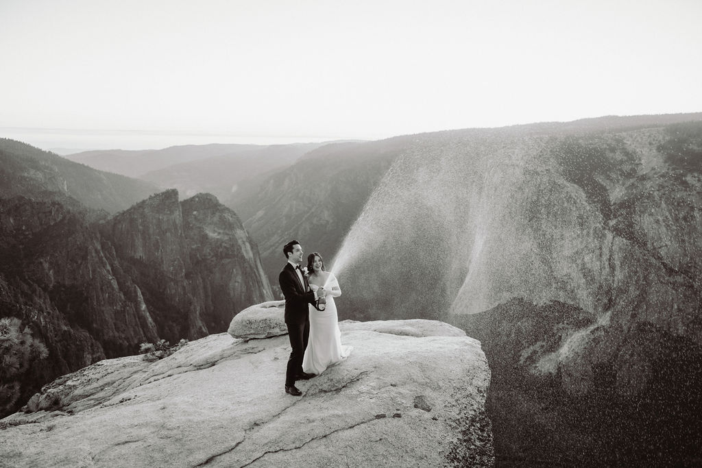 A bride and groom stand on a cliff edge, with the groom spraying champagne while the bride smiles, set against a backdrop of mountains and a sunset sky.