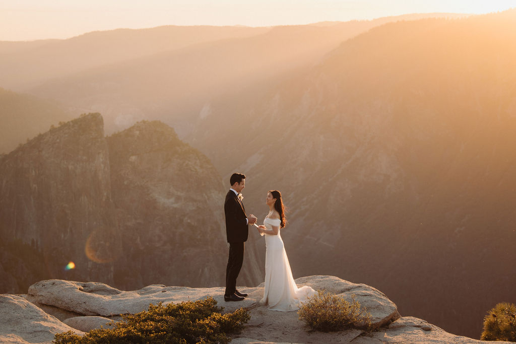 A couple stands facing each other at the edge of a cliff during sunset. The man is in a suit, and the woman is in a white dress, with mountains in the background at Taft Point 